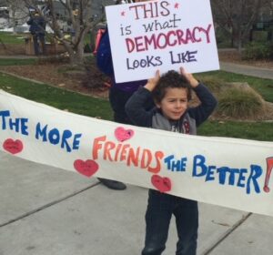 A child holds a sign saying, "This is what democracy looks like" and a banner says, "The more friends the better!"